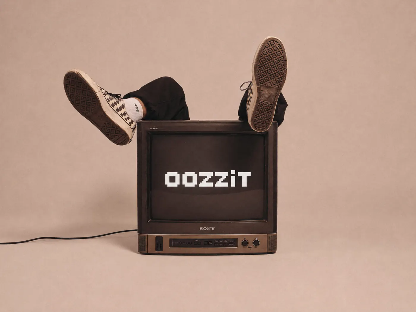 A CRT television showing the Oozzit wordmark, with a pair of legs in checkered sneakers kicked up behind it.